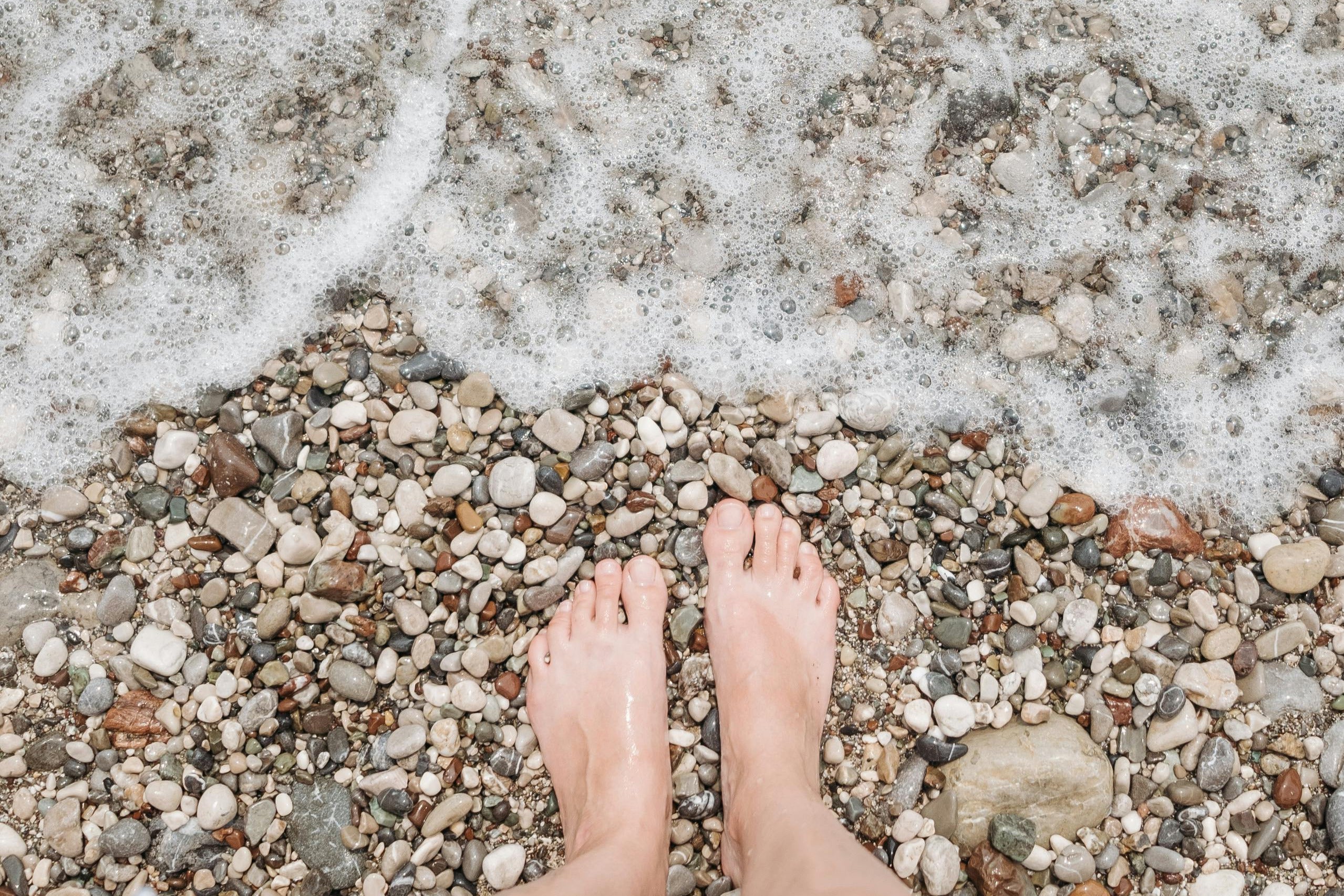Feet standing on a pebbly shore with waves crashing onto the shore.