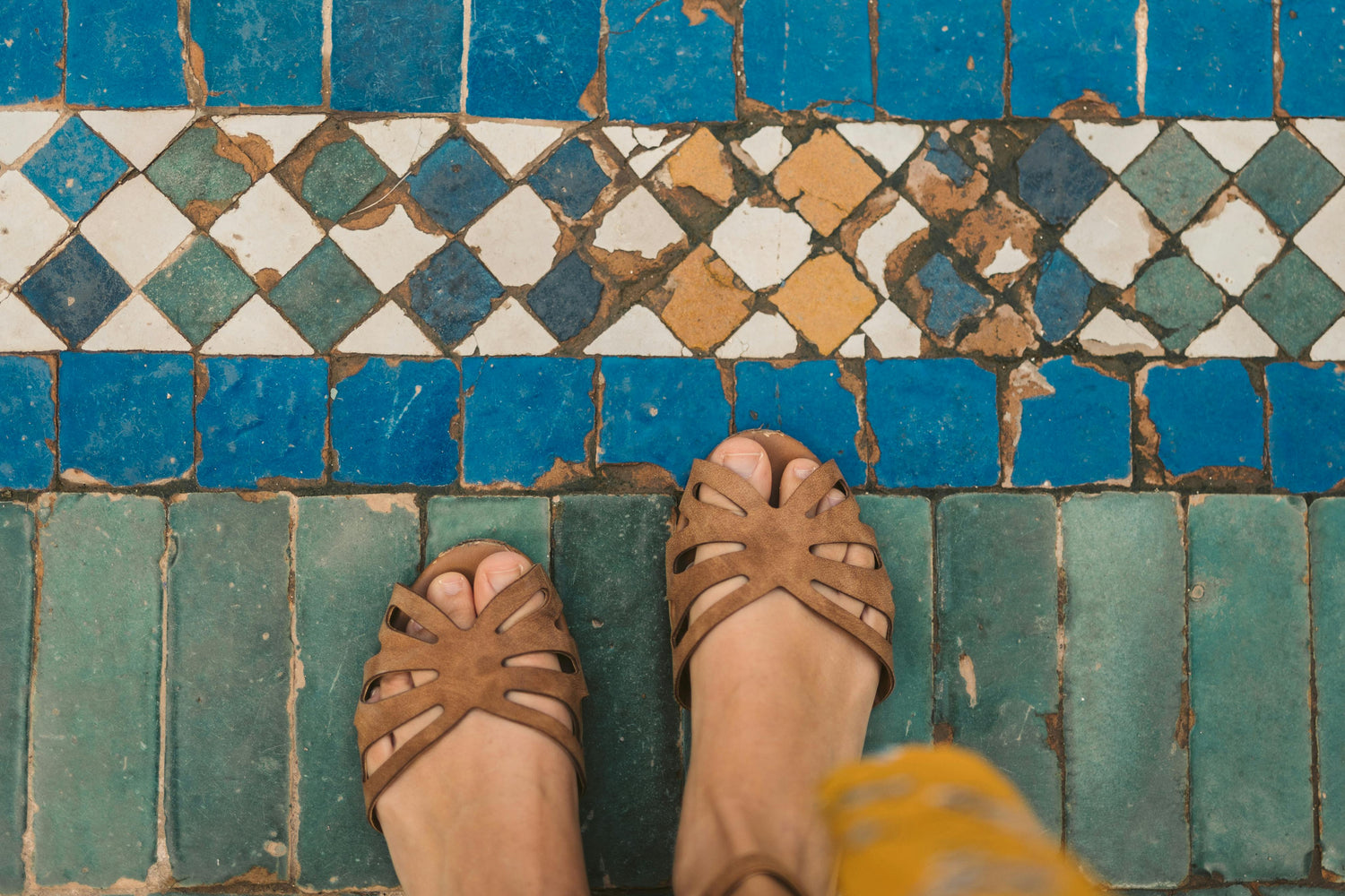 Feet wearing brown sandals on a mosaic tile floor with blue and green tiles.